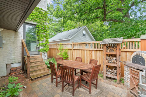 A cozy outdoor patio featuring a wooden dining table with chairs, a staircase, and a wood storage rack surrounded by trees.