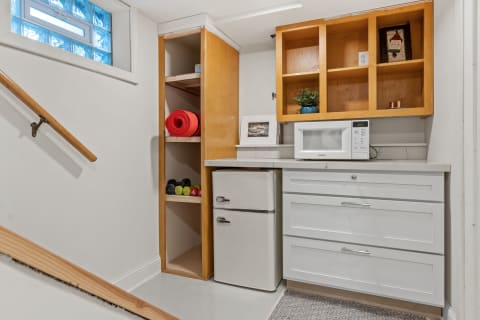 A bright basement kitchenette featuring a fridge, microwave, and exercise equipment.