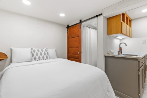 A modern bedroom featuring a bed with white linens, a decorative pillow, sliding wooden door, and a small kitchenette.
