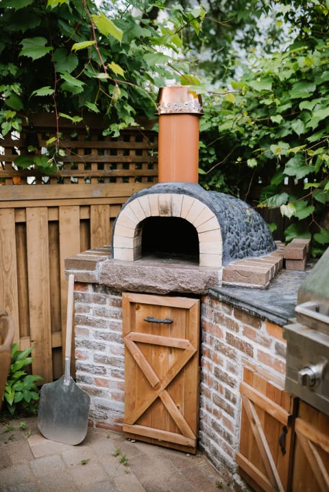 Outdoor brick oven with a copper chimney and wooden cabinet, set in a green garden.