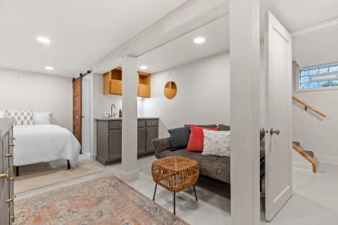 A cozy interior space featuring a bed, a dark sofa with colorful pillows, a side table, and a kitchenette in a basement setting.