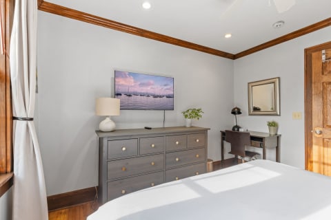 A cozy bedroom featuring a dresser, TV, desk, and natural light from a window.