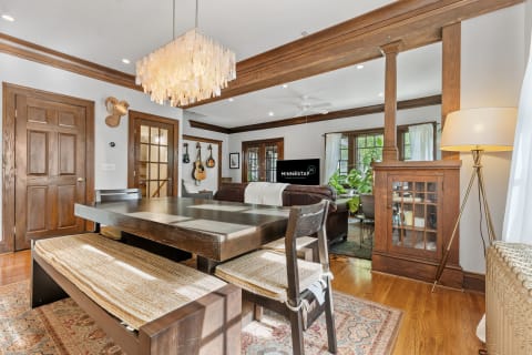 A modern dining area featuring a wooden table, bench seating, and chandelier, leading into a living room with plants and a leather sofa.