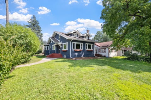 Blue craftsman home with a spacious lawn and trees in a residential area under a blue sky.