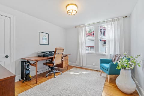 Bright home office with a wooden desk, ergonomic chair, teal armchair, and natural light from large windows.