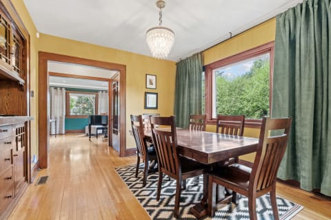 Cozy dining room featuring warm wooden furniture, a chandelier, and large windows with green curtains.