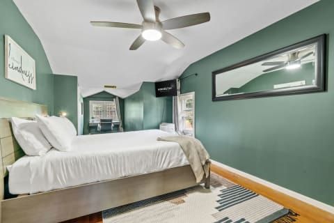 A sage-green bedroom with a bed, wall-mounted TV, and work desk by the window.