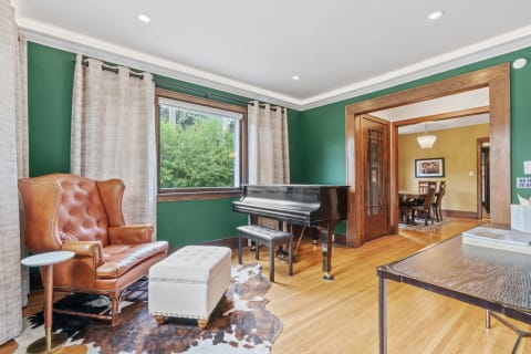 A luxurious living room with a leather armchair, grand piano, and window, featuring green walls and hardwood floors.