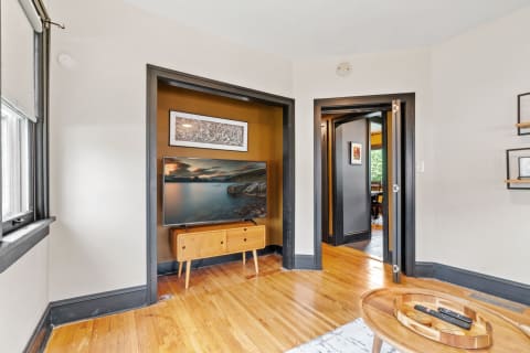 A cozy living area with a television on a wooden console, hardwood flooring, and a doorway leading to another room.