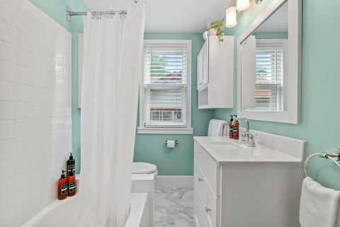 A serene bathroom featuring mint walls, white fixtures, and marble floors, showcasing a bathtub with a shower curtain and a sleek vanity.