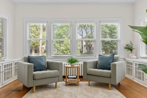 A bright sitting area featuring two grey armchairs with blue cushions, a small wooden table, and windows showing greenery outside.