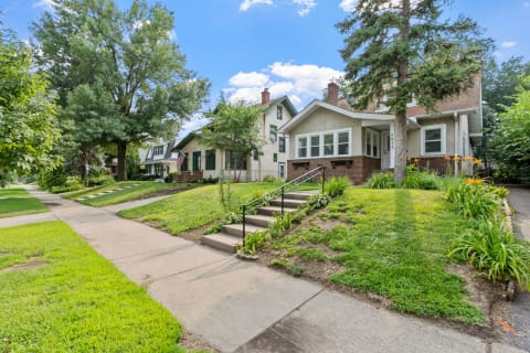 A residential street scene featuring two homes with green lawns and gardens under a blue sky.