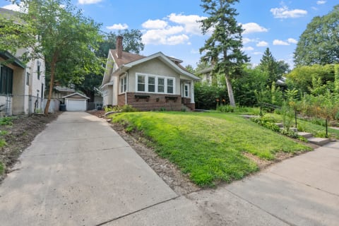 A traditional-style house with a green lawn and a driveway, surrounded by trees and flowers.
