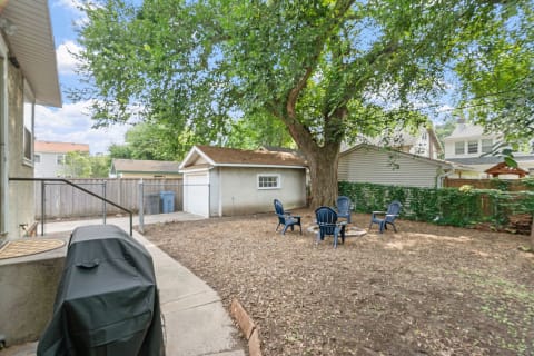 Backyard with a large tree, blue chairs around a fire pit, and a garage in the background.