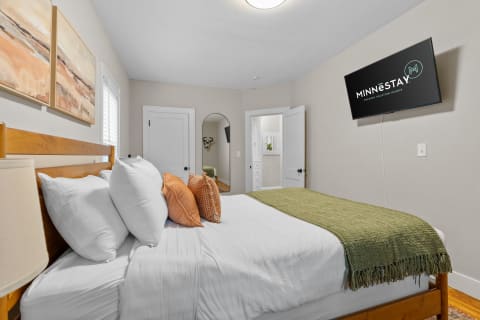 Cozy bedroom featuring a wooden bed with white bedding and decorative pillows, a wall-mounted TV, and abstract artwork.