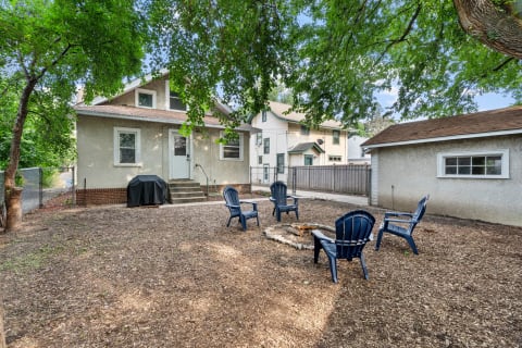 Backyard with a fire pit and blue chairs next to a house and a tree.