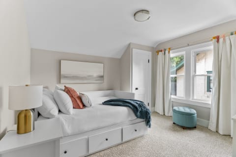 A modern bedroom with a white daybed, elegant lamp, and bright windows.