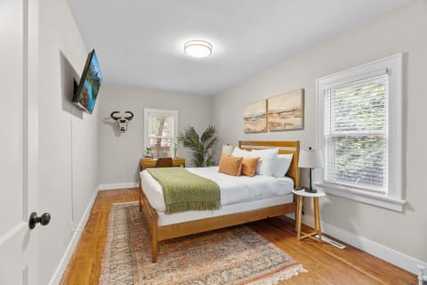 Cozy bedroom with wooden furniture and soft colors, including a bed with decorative pillows and a desk by the window.