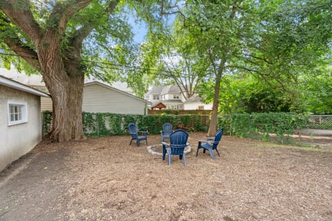 Backyard scene featuring a stone fire pit surrounded by blue chairs and a large tree.