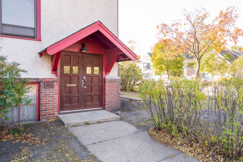 A building entrance with brown double doors and a red overhang, surrounded by autumn plants.