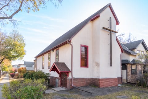 A charming church building with red-framed windows and a wooden cross, surrounded by autumn foliage.