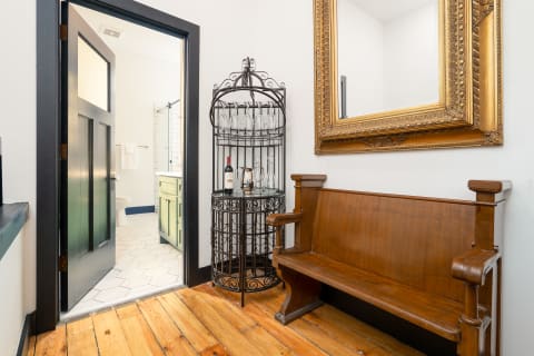 A cozy entryway with a wooden bench, wrought iron bar cart, and a view of a bathroom.