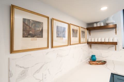 Modern kitchen corner with framed artwork and wooden shelves displaying white cups.