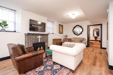 A living room featuring a white tufted sofa, a brown leather armchair, and a modern fireplace with a TV above it.