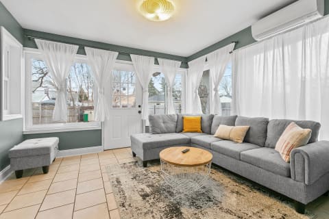 A sunroom with a gray sectional sofa, large windows, and a wooden coffee table.