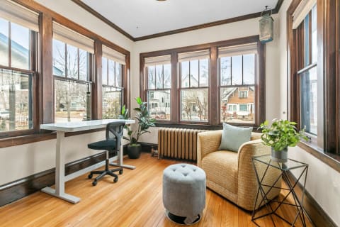 A sunroom workspace with large windows, wooden floors, a modern desk, an armchair, and a potted plant.