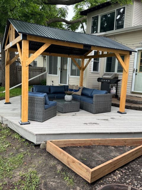 An outdoor deck with a pergola, featuring navy blue cushions and a glass table, alongside a grill and a planter box.