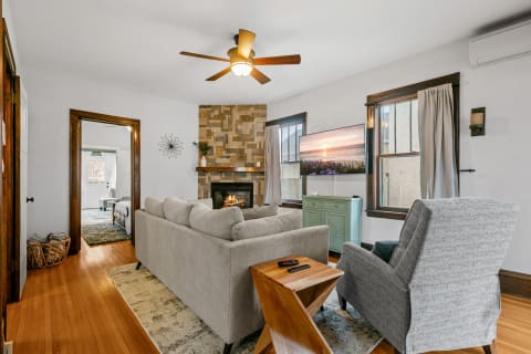 Cozy living room with gray sectional sofa, stone fireplace, and a view of a neighboring room.
