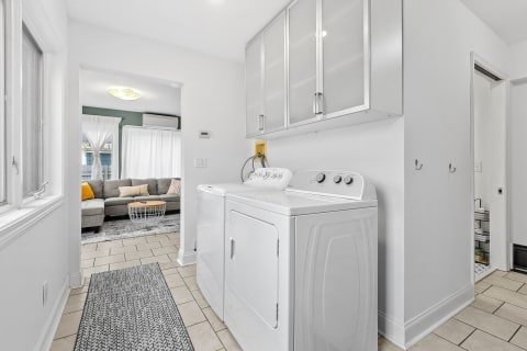 Bright laundry room showcasing a full-size washer and dryer with modern cabinets and a glimpse of a cozy living room.