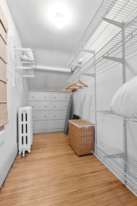 A spacious walk-in closet featuring white shelving, wooden hangers, a vintage radiator, and natural light illuminating the wooden floor.