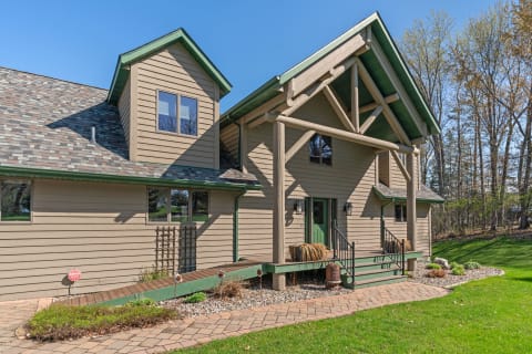 A picturesque home featuring light brown siding, green trim, and a slate roof with a welcoming entrance and landscaped yard.