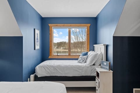 A bedroom with blue walls, two beds, and a large window overlooking trees and a river.