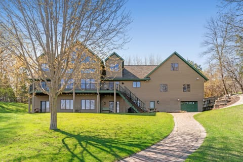 A large two-story house with brown siding and green trim, featuring a deck and numerous large windows, set against a blue sky, with a paved path leading to it.