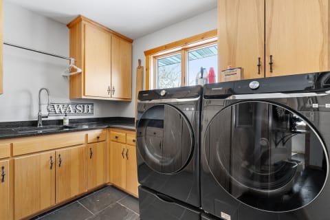 Interior of a modern laundry room featuring Samsung washing machines, wooden cabinets, and a sink.