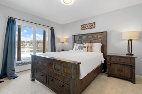 Cozy bedroom featuring a wooden bed, decorative pillows, and a view of a lake through large windows.