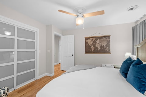 A cozy bedroom featuring a bed with white bedding and teal pillows, a world map on the wall, and a ceiling fan.