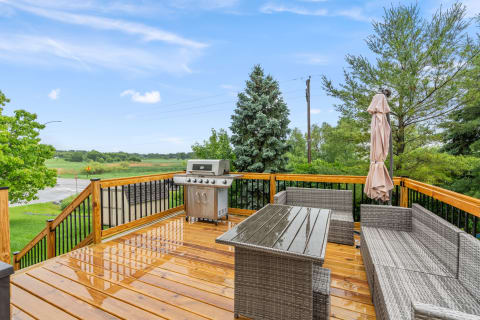 A wooden deck featuring wicker furniture and a grill, with a scenic view of green fields and a blue sky.