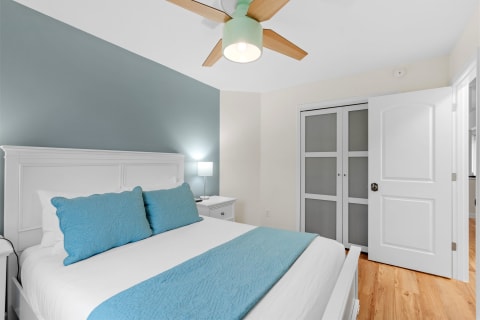 A peaceful bedroom featuring a white bed with blue pillows, a ceiling fan, and wooden flooring.