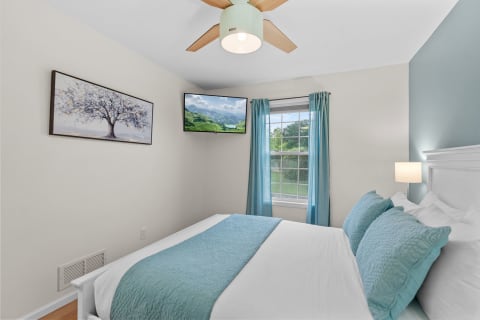 A cozy bedroom featuring a comfortable bed with blue accents, a mounted TV, and a window with turquoise curtains.
