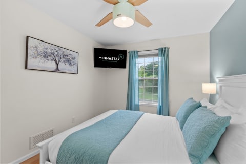 A modern bedroom with a white bed and teal pillows, light curtains, a ceiling fan, and a wall-mounted TV.