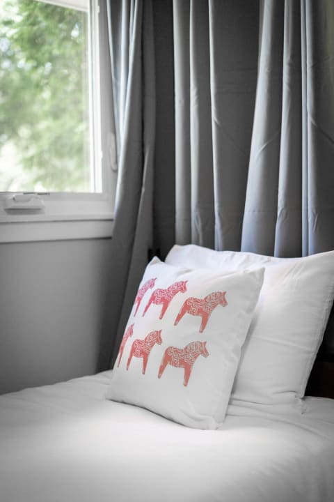 A decorative pillow with elephant designs on a neatly made white bed with gray curtains and a window letting in light.
