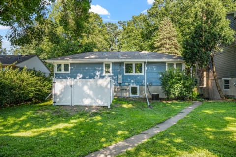 A single-story house with light blue siding and a white fence, set in a green yard with trees and a clear blue sky.