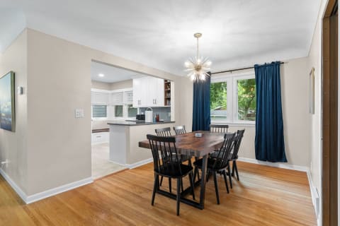 A dining area with a wooden table surrounded by black chairs and a bright chandelier, leading to a kitchen.