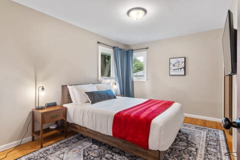 A cozy bedroom featuring a queen-sized bed with white bedding and a red throw blanket, a nightstand with a lamp, and blue curtains.