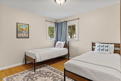 A cozy bedroom featuring two single beds, decorative pillows, a colorful landscape painting, and natural light.