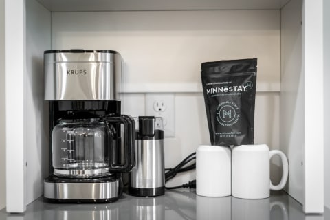 A KRUPS coffee maker next to a coffee grinder and MINNESTAY coffee grounds in a cabinet.